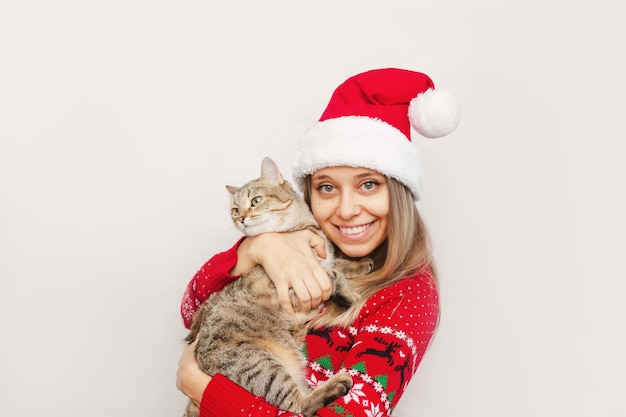 Una mujer joven en un suéter de ciervo rojo gorro de Papá Noel sosteniendo un gato atigrado Navidad Año Nuevo conep