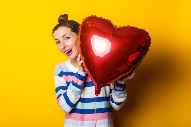 Mujer joven con un suéter se esconde detrás de un corazón de globo de aire sobre un fondo amarillo. Composición del día de San Valentín.