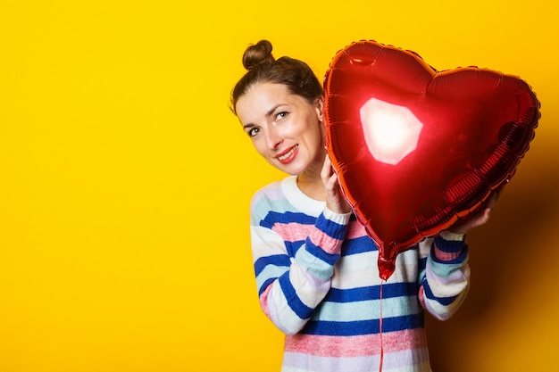 Mujer joven con un suéter se esconde detrás de un corazón de globo de aire sobre un fondo amarillo. Composición del día de San Valentín.