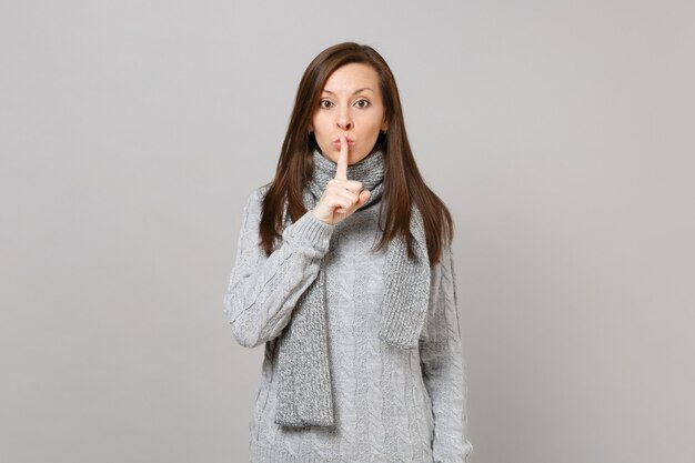 Mujer joven en suéter gris, bufanda diciendo silencio, cállate con el dedo en los labios, gesto shhh aislado sobre fondo gris. Concepto de estación fría de emociones de estilo de vida de moda saludable. Simulacros de espacio de copia.