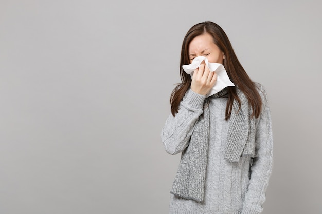 Mujer joven en suéter gris, bufanda con secreción nasal, sonarse la nariz a la servilleta aislado sobre fondo de pared gris. Estilo de vida saludable, tratamiento de enfermedades enfermas, concepto de estación fría. Simulacros de espacio de copia.