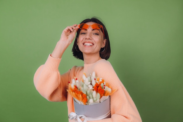 Mujer joven en suéter de melocotón casual aislado en la pared de olivo verde sostenga la composición de la caja de flor blanca naranja de flores de algodón, gypsophila, trigo y lagurus para un regalo feliz sorprendido