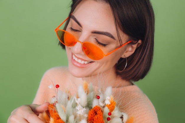 Mujer joven en suéter de melocotón casual aislado en la pared de olivo verde sostenga la composición de la caja de flor blanca naranja de flores de algodón, gypsophila, trigo y lagurus para un regalo feliz sorprendido