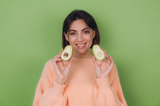 Mujer joven en suéter de melocotón casual aislado en la pared verde oliva con aguacate, concepto de salud y cuidado de la piel, espacio de copia