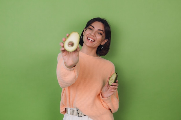 Mujer joven en suéter de melocotón casual aislado en la pared verde oliva con aguacate, concepto de salud y cuidado de la piel, espacio de copia