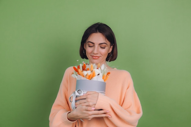 Mujer joven en suéter de melocotón casual aislado en la pared verde oliva sostenga la composición de la caja de flor blanca naranja de flores de algodón, trigo de gypsophila y lagurus para un regalo feliz sorprendido