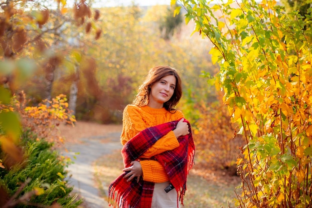 Una mujer joven con un suéter naranja se envuelve en una bufanda en un parque de otoño