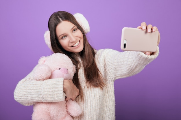 Una mujer joven con un suéter y orejas rosas se toma una selfie y la abraza con un juguete.