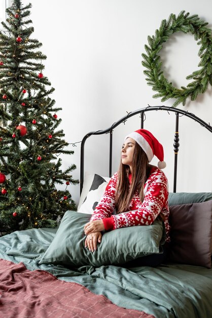 Mujer joven en suéter rojo y gorro de Papá Noel sentado en la cama celebrando la Navidad