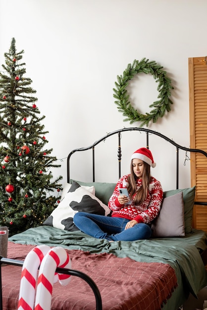 Mujer joven en suéter rojo y gorro de Papá Noel sentado en la cama con mensajes de texto de teléfono móvil o videollamada