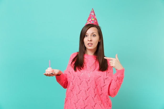 Mujer joven en suéter rosa de punto, sombrero de cumpleaños que señala el dedo índice en la torta con vela en mano aislado en retrato de estudio de fondo azul turquesa. Concepto de estilo de vida de personas. Simulacros de espacio de copia.