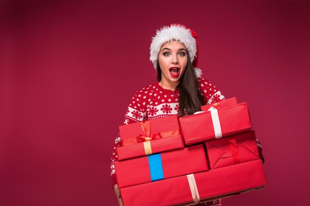 Mujer joven en suéter y sombrero de navidad sosteniendo cajas de regalo y mirando a la cámara sobre fondo rojo.