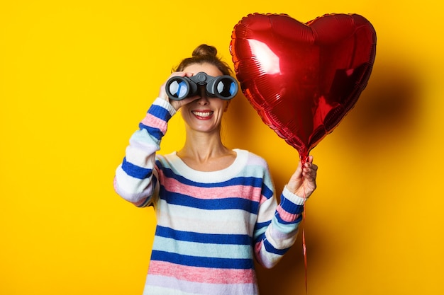 Mujer joven con un suéter sostiene un corazón de globo de aire y mira a través de binoculares sobre un fondo amarillo. Composición del día de San Valentín.