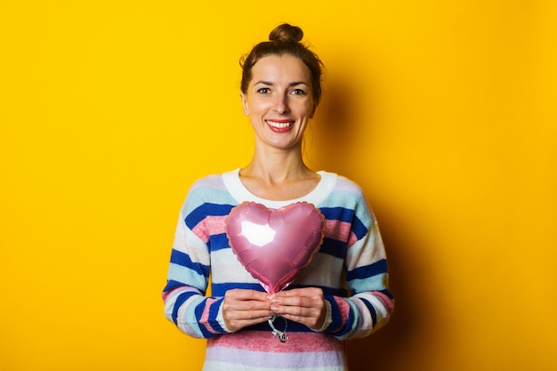 Mujer joven con un suéter sostiene un corazón de globo de aire sobre un fondo amarillo. Composición del día de San Valentín.