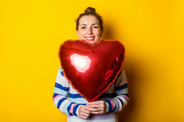 Mujer joven con un suéter sostiene un corazón de globo de aire sobre un fondo amarillo. Composición del día de San Valentín.