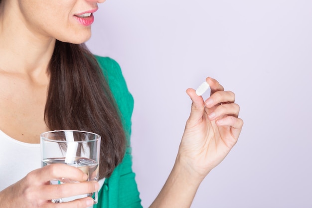 Mujer joven en suéter verde y camisa blanca con una píldora y un vaso de agua. Concepto de medicina y salud.