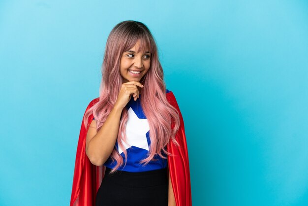 Mujer joven superhéroe con cabello rosado aislado sobre fondo azul mirando hacia el lado y sonriendo