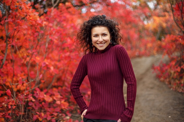 Mujer joven en sweather rojo posando en la cámara en otoño rojo arbustos