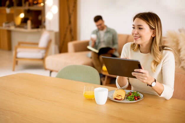 Mujer joven con tableta digital y desayunando en la cocina con un novio sentado en el fondo