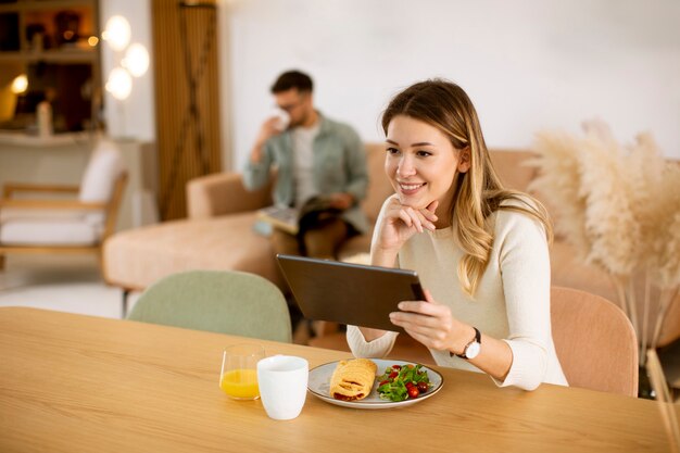 Mujer joven con tableta digital y desayunando en la cocina con un novio sentado