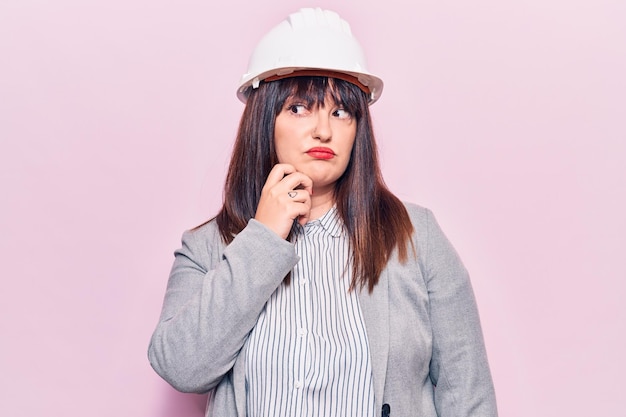 Mujer joven de talla grande con casco de arquitecto pensando concentrada en la duda con el dedo en la barbilla y mirando hacia arriba preguntándose