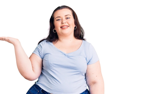 Mujer joven de talla grande con ropa informal sonriendo alegre presentando y señalando con la palma de la mano mirando a la cámara