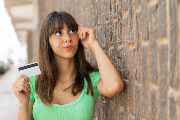 Mujer joven con una tarjeta de crédito al aire libre que tiene dudas y con una expresión facial confusa