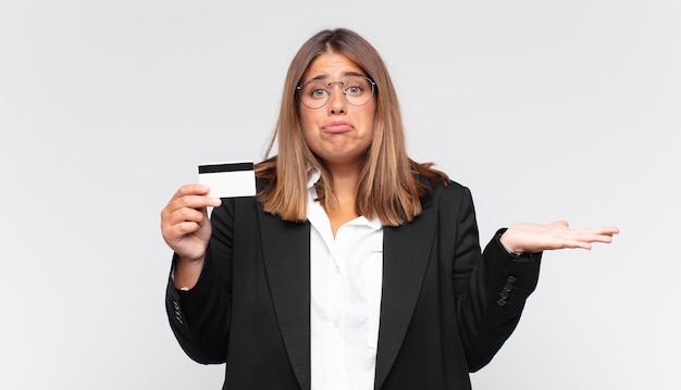 Mujer joven con una tarjeta de crédito que se siente perpleja y confundida, dudando, ponderando o eligiendo diferentes opciones con expresión divertida