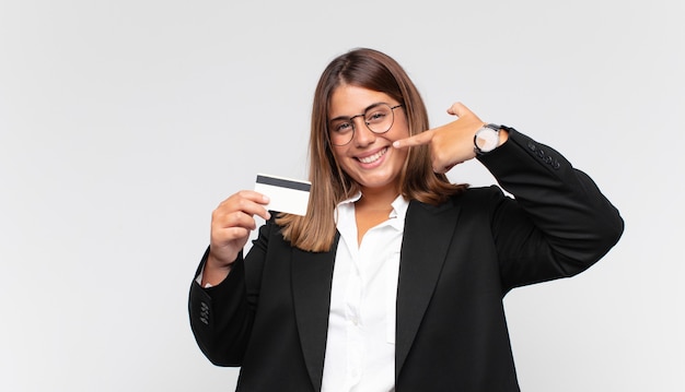Mujer joven con una tarjeta de crédito sonriendo con confianza apuntando a su propia sonrisa amplia, actitud positiva, relajada y satisfecha