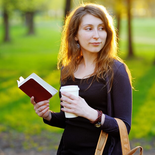 Mujer joven con una taza de café y un libro en el parque al atardecer