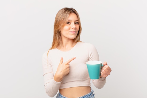 Mujer joven con una taza de café que parece emocionada y sorprendida apuntando hacia un lado