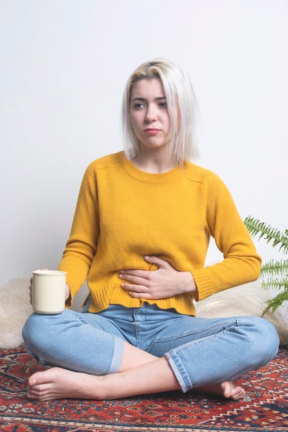 Mujer joven con taza de café sufriendo de dolor de estómago contra la pared en casa