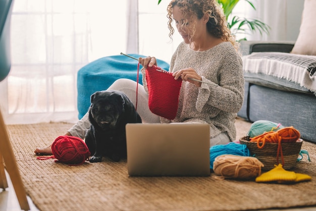 Mujer joven tejiendo lana con aguja mientras ve el tutorial en línea en la computadora portátil y se sienta en el piso con su perro en casa. Mujer viendo lecciones de costura, una computadora portátil y pasatiempos en casa.
