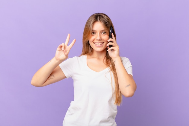 Mujer joven con un teléfono celular sonriendo y mirando amigable, mostrando el número dos
