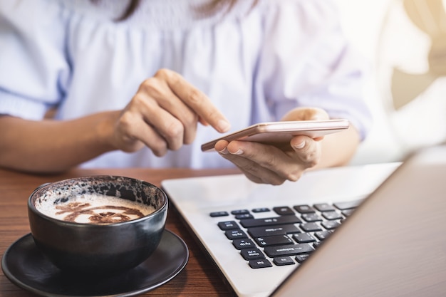 Mujer joven con teléfono inteligente y computadora portátil con una taza de café en la cafetería
