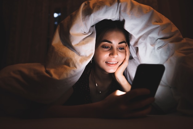 Mujer joven con un teléfono inteligente debajo de una manta en una cama