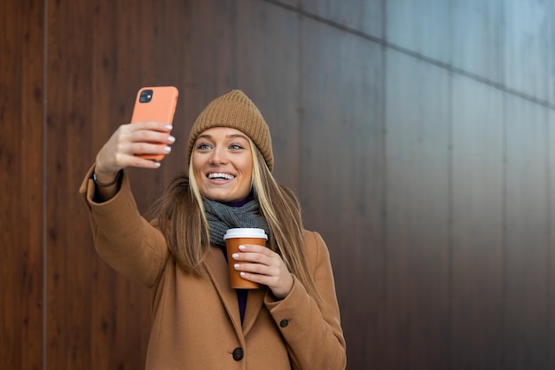 Mujer joven con teléfono inteligente de pie contra la calle fondo de edificio borroso Foto de moda de una chica hermosa con teléfono y taza de café