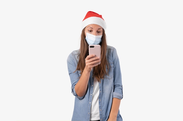 Mujer joven con teléfono, máscara médica y gorro de Papá Noel. Compras navideñas online en cuarentena.