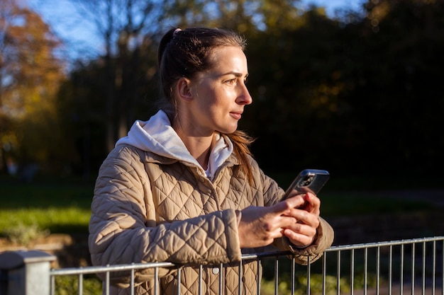 Mujer joven con un teléfono mira hacia otro lado en el fondo de un parque de otoño