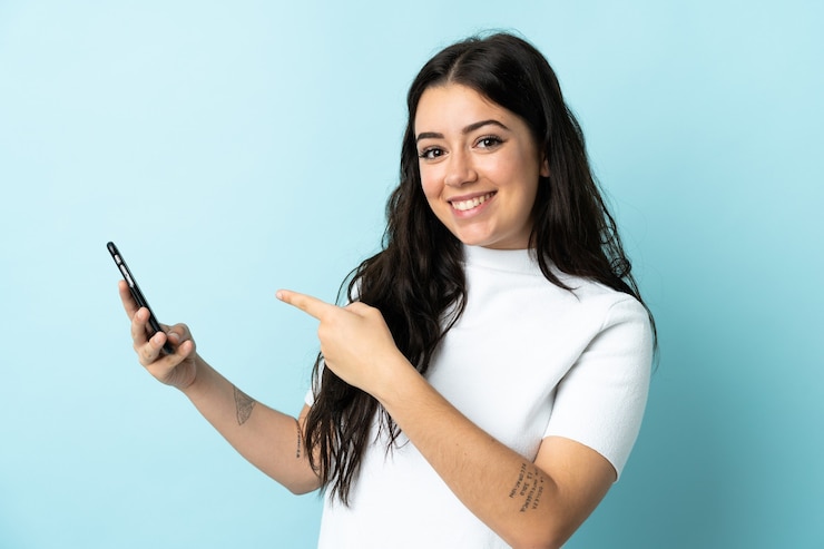 Foto mujer joven con teléfono móvil aislado en la pared azul y apuntando