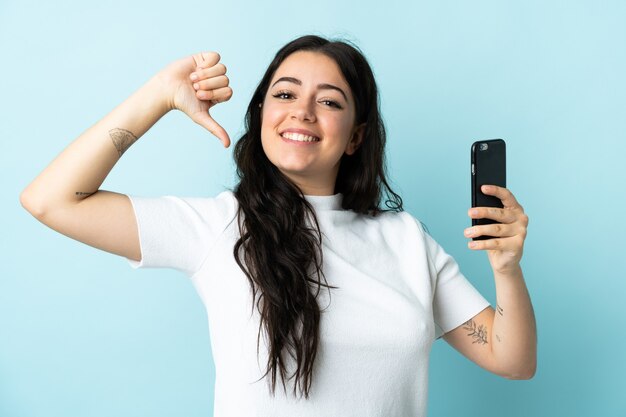 Mujer joven con teléfono móvil aislado en la pared azul orgulloso y satisfecho de sí mismo