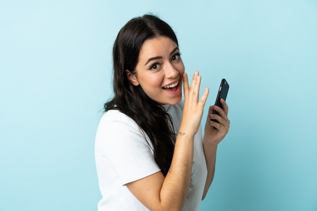 Mujer joven con teléfono móvil aislado sobre fondo azul susurrando algo