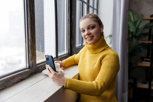 Mujer joven con teléfono móvil y auriculares mientras se queda en casa con una taza de café