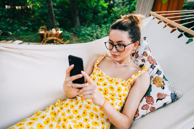 Mujer joven con teléfono móvil, relajándose en una hamaca al aire libre, en el jardín trasero.