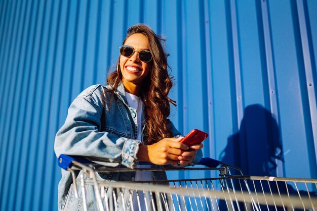 Mujer joven con teléfono sentado carrito de compras divirtiéndose cerca de la pared de color Cliente adicto a las compras