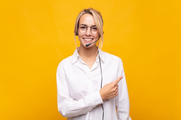 Mujer joven telemarketer sonriendo alegremente, sintiéndose feliz y apuntando hacia un lado y hacia arriba, mostrando el objeto en el espacio de copia