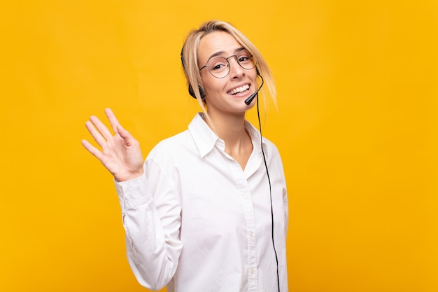 Mujer joven telemarketer sonriendo feliz y alegremente, saludando con la mano, dándole la bienvenida y saludándolo, o despidiéndose