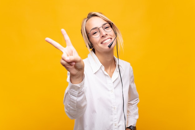 Mujer joven telemarketer sonriendo y mirando feliz, despreocupado y positivo, gesticulando victoria o paz con una mano