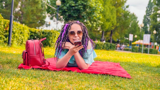 Mujer joven tendida en una manta graba un mensaje de voz usando un teléfono inteligente en el parque Niña feliz tendida en el césped y usa la aplicación de grabación de voz o dicta un mensaje de audio