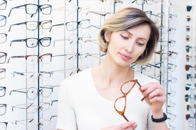 Mujer joven en tienda de óptica eligiendo nuevas gafas con gafas ópticas en la tienda de óptica Una mujer elige gafas Emociones Oftalmología